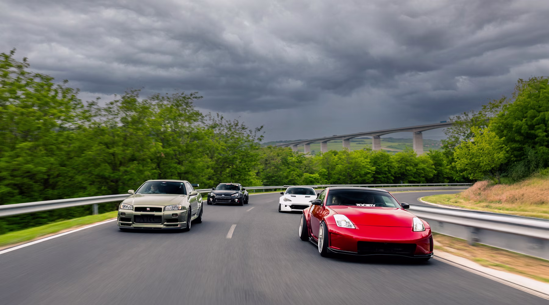 Several cars driving in convoy on a winding road under stormy skies